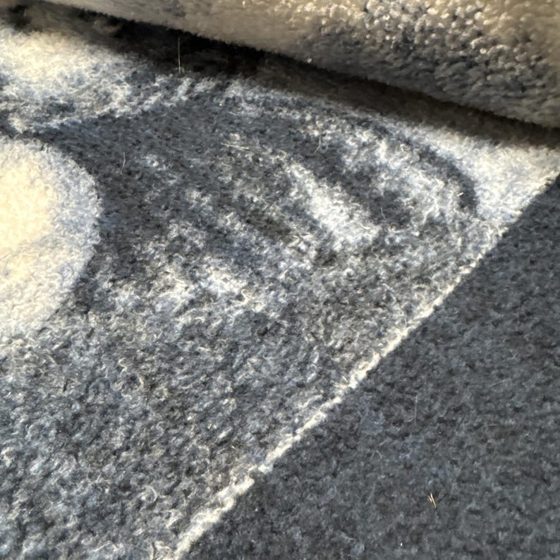 Puede incluir: Primer plano de una alfombra enrollada con una pila texturizada y en bucle. La alfombra presenta un patr&oacute;n de formas grises claras y blancas sobre un fondo gris m&aacute;s oscuro. El borde de la alfombra es visible, mostrando la parte enrollada.