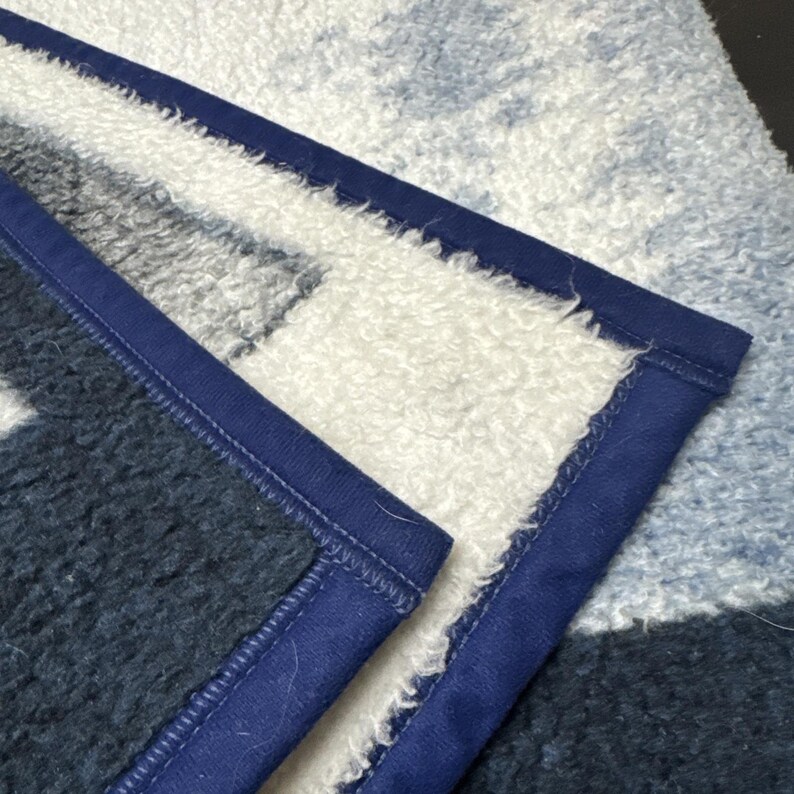 Puede incluir: Primer plano de una manta suave y afelpada con un patr&oacute;n geom&eacute;trico en tonos de azul, blanco y gris. La manta tiene un borde azul oscuro y una superficie texturizada y esponjosa.