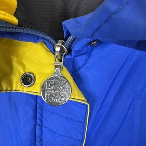 May include: Close-up of a blue and yellow jacket with a silver zip. The zip pull has a circular emblem with the word "BOMBARDIER" on it. The jacket has a black lining and a snap closure.