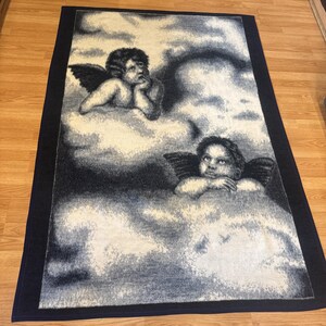 Puede incluir: Una alfombra rectangular con un dise&ntilde;o celestial con dos figuras de querubines con alas descansando sobre nubes. La alfombra tiene un borde azul marino y una paleta de colores blanco, gris y negro.