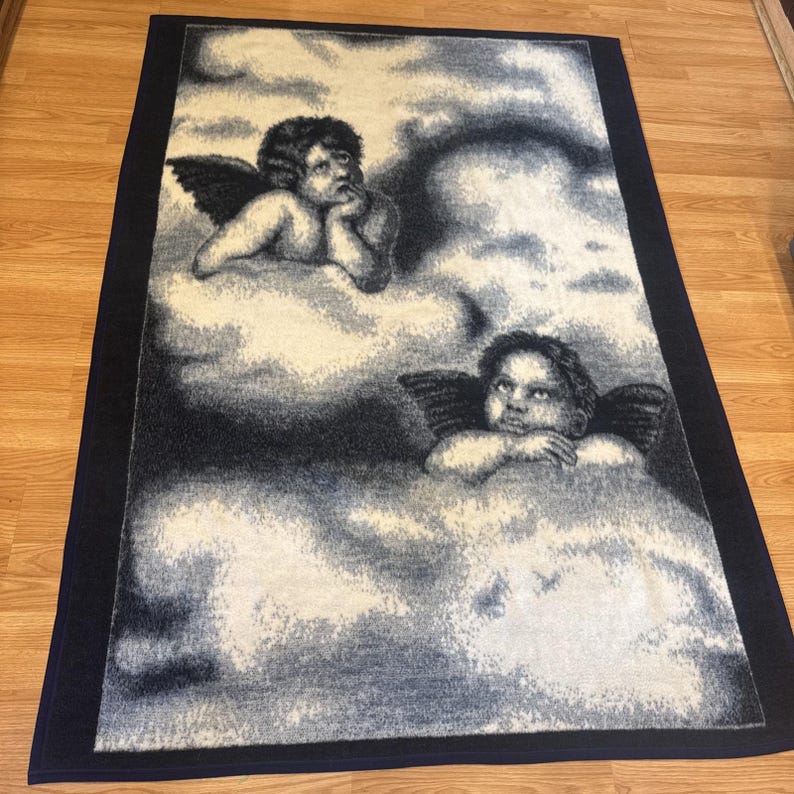 Puede incluir: Una alfombra rectangular con un dise&ntilde;o celestial con dos figuras de querubines con alas descansando sobre nubes. La alfombra tiene un borde azul marino y una paleta de colores blanco, gris y negro.