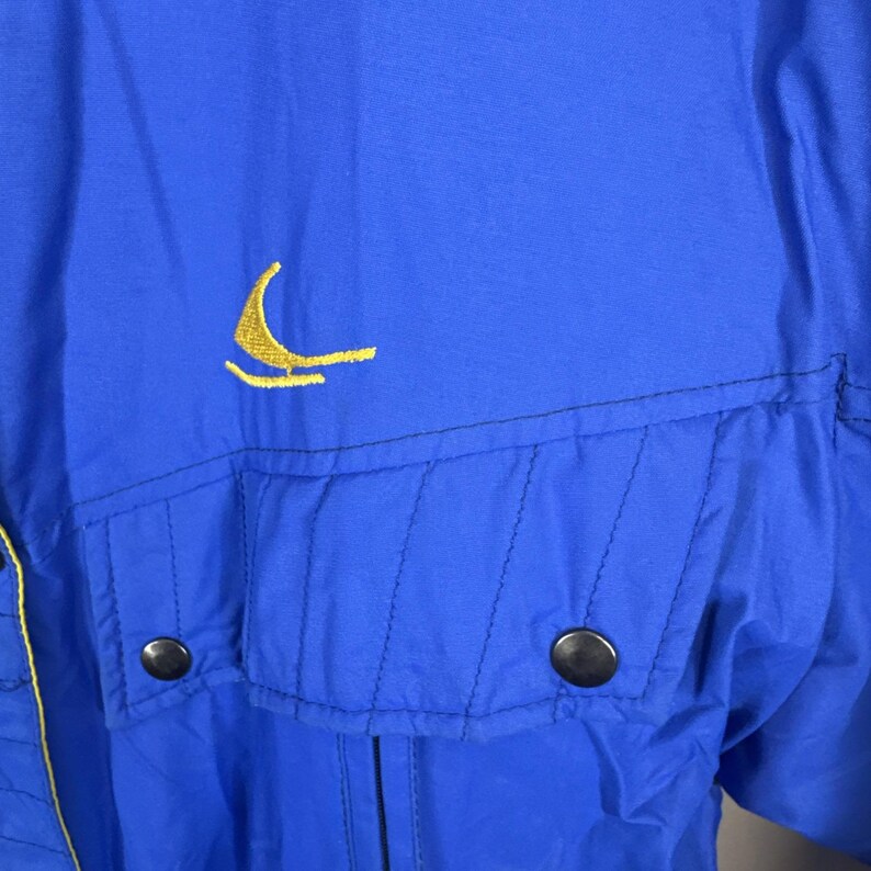 May include: Close-up of a blue jacket featuring a yellow embroidered sailboat logo. The jacket has two front pockets with black button closures and yellow trim. The fabric appears to be a lightweight nylon material.