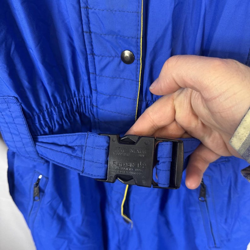 May include: Close-up of a blue snowsuit with a black buckle and a yellow accent. The snowsuit has a cinched waist and a zipper pocket. The buckle has the text "TW Nexus" and "Ender-1".