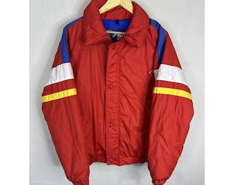 May include: A red vintage ski jacket with blue and white accents. The jacket has a high collar, snap closures, and yellow stripes on the sleeves. It is hanging on a wooden hanger against a white background.