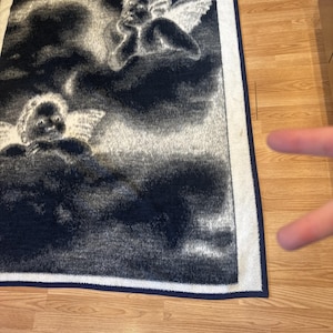 Puede incluir: Una manta rectangular azul oscuro y blanca con un dise&ntilde;o celestial con dos &aacute;ngeles querubines entre nubes. La manta tiene un borde blanco con un ribete azul oscuro.