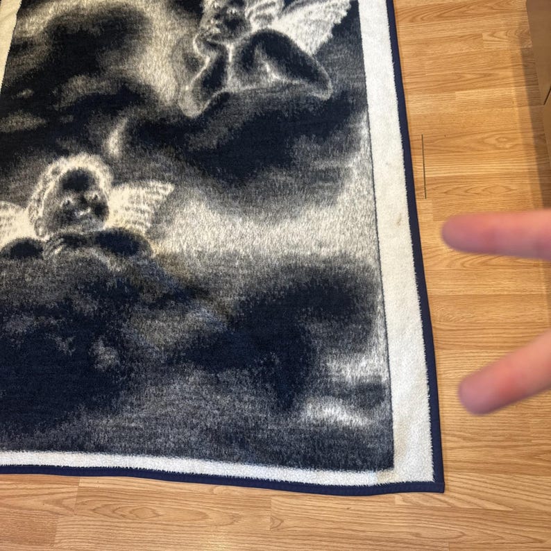 Puede incluir: Una manta rectangular azul oscuro y blanca con un dise&ntilde;o celestial con dos &aacute;ngeles querubines entre nubes. La manta tiene un borde blanco con un ribete azul oscuro.