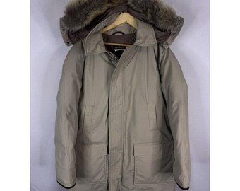 May include: A tan winter parka with a fur-trimmed hood. The coat has a button and zipper closure, two front pockets, and a brown lining. The parka is hanging on a wooden hanger.