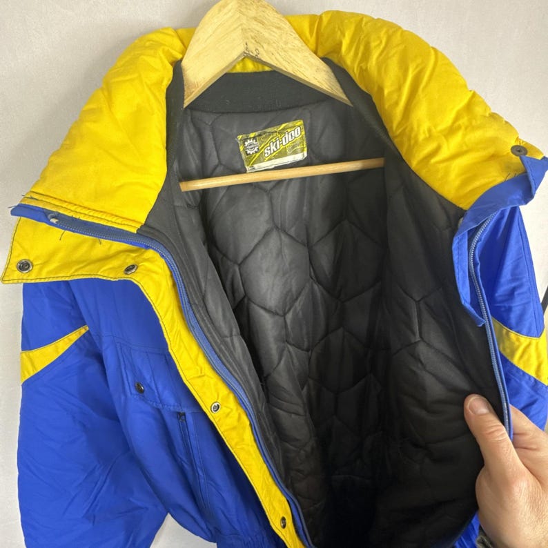 May include: A vintage ski jacket in blue, yellow, and black. The jacket has a high collar, a quilted black lining, and the "Ski-doo" brand label. The jacket is hanging on a wooden hanger.