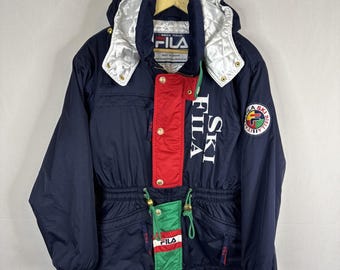 May include: Navy blue FILA ski jacket with a red and green accent panel. The jacket features the FILA logo and "SKI FILA" in white lettering. It has a hood with silver lining and gold buttons.