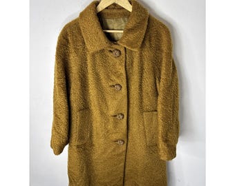 Peut inclure: Un manteau vintage, mi-long, de couleur brun doré. Le manteau présente un tissu texturé et duveteux, un large col et trois gros boutons ronds sur le devant. Il possède deux poches avant et est suspendu à un cintre en bois.