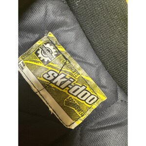 May include: Close-up of a black garment with a yellow and white Ski-doo Sportswear label. The label features the Bombardier logo and text. The garment appears to be made of a durable, textured material.