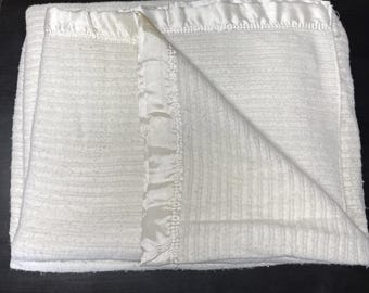 Peut inclure: Une couverture blanche avec une surface texturée et côtelée, bordée d'une finition en satin lisse et brillant. La couverture est pliée pour révéler la texture intérieure et les bords finis du tissu, présentant une teinte blanche uniforme.
