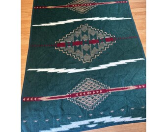 Peut inclure: Une couverture verte avec un motif géométrique. La couverture présente des formes de losanges dans des tons de marron, de rouge et de beige, avec des rayures horizontales blanches et rouges. La couverture est rectangulaire et semble être faite d'une matière douce.
