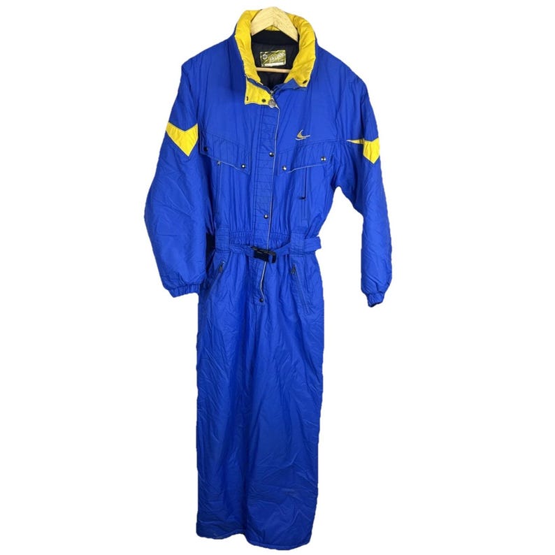 May include: A blue and yellow ski suit. The one-piece suit features a high collar, a belt at the waist, and yellow accents on the shoulders and sleeves. The suit is made of a water-resistant material, designed for cold weather activities.