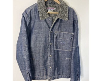 May include: A dark blue denim jacket with a sherpa collar and a single chest pocket. The jacket has a button closure and long sleeves.