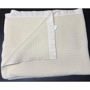 May include: A cream-colored thermal blanket featuring a textured, waffle-knit surface and a smooth, matching satin trim along the edges. The blanket is folded over at one corner to reveal the consistent weave and the satin binding detail.