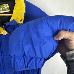 May include: A blue and yellow Ski-Doo winter jacket. The jacket has a black collar and a yellow hood. The sleeve is puffed out, showing the quilted design. The jacket has a zipper and a Ski-Doo logo patch.