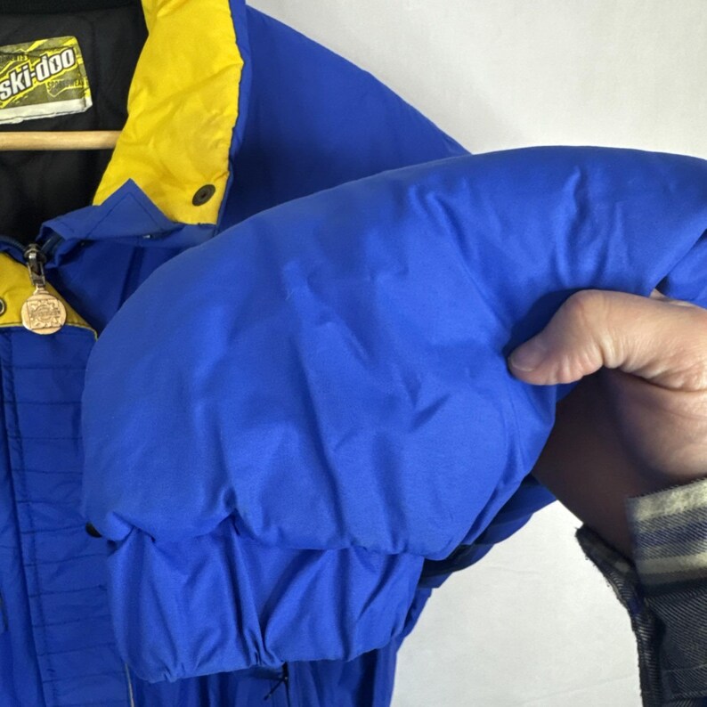 May include: A blue and yellow Ski-Doo winter jacket. The jacket has a black collar and a yellow hood. The sleeve is puffed out, showing the quilted design. The jacket has a zipper and a Ski-Doo logo patch.