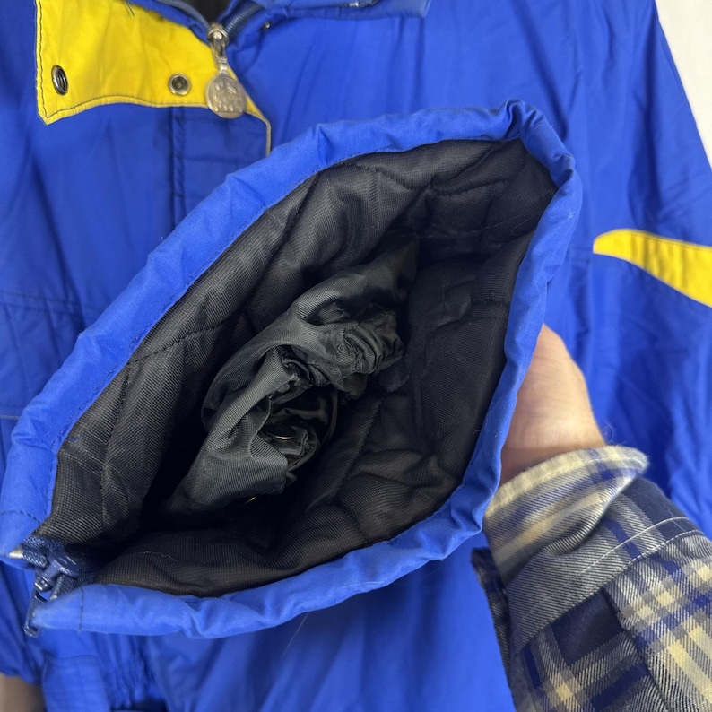 May include: A blue and yellow winter jacket with a black quilted lining. The jacket has a zip closure and a high collar. The sleeve is open, revealing the black lining and a drawstring cuff.