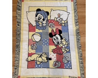 Peut inclure: Une couverture Disney rectangulaire pour bébé avec Mickey Mouse, Minnie Mouse et Pluto. La couverture a un motif patchwork avec des carrés rouges, bleus et jaunes. La couverture a une bordure à franges et le logo Disney.