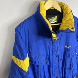 May include: A blue and yellow ski jacket with the brand name "Ski Doo" on the label. The jacket has a high collar, a zipper closure, and multiple pockets. The jacket has yellow accents on the shoulders and collar.
