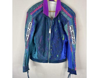 May include: A vintage Phenix ski jacket with a vibrant color scheme. The jacket features a purple collar, a blue and teal body with colorful paint splatter design, and the Phenix logo in white on the sleeves. It has a full zipper and drawstrings.