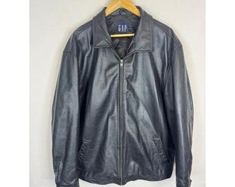 Peut inclure: Veste en cuir noire avec fermeture éclair, deux poches avant et un col. La veste est suspendue à un cintre en bois. Le nom de la marque "GAP" est visible sur l'étiquette.
