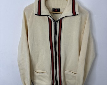 Peut inclure: Un pull zippé en tricot crème avec des rayures rouges et noires sur le devant. Le pull a deux poches et un col montant.