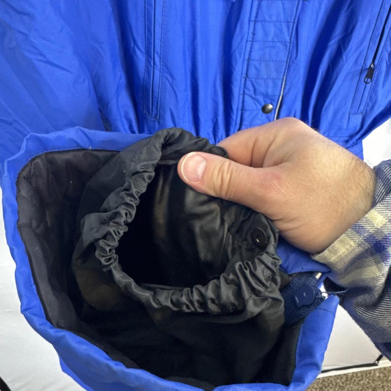 May include: A bright blue jacket with a black interior sleeve and elastic cuff. The jacket has a button closure and a zipper detail. The sleeve is being held open, revealing the black interior. The jacket appears to be made of a lightweight material.