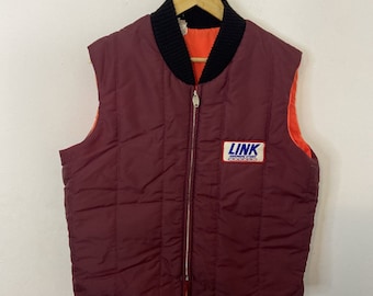 Peut inclure: Un gilet matelassé marron avec un col côtelé noir et une fermeture éclair. Le gilet a un patch sur le devant qui dit "LINK AUTOMOTIVE STORES".