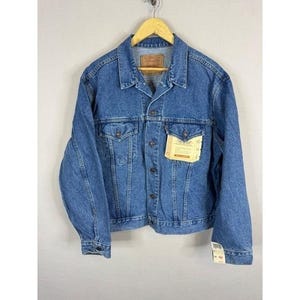 May include: A medium-wash denim jacket with a classic collar, button-down front, and two chest pockets. The jacket has a hang tag attached and is displayed on a wooden hanger. The jacket is a size M.