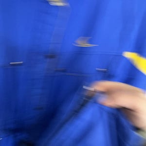 May include: A bright blue jacket with a yellow detail. The jacket features buttons and a pocket. The image is slightly blurred, but the colour and style of the jacket are evident.