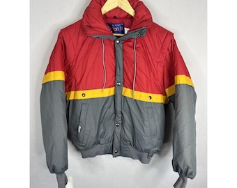 May include: A vintage red, yellow, and gray winter jacket with a high collar. The jacket has a color-blocked design with a red upper section, yellow stripe, and gray sleeves and lower body. It features a button-up front and a small tag.