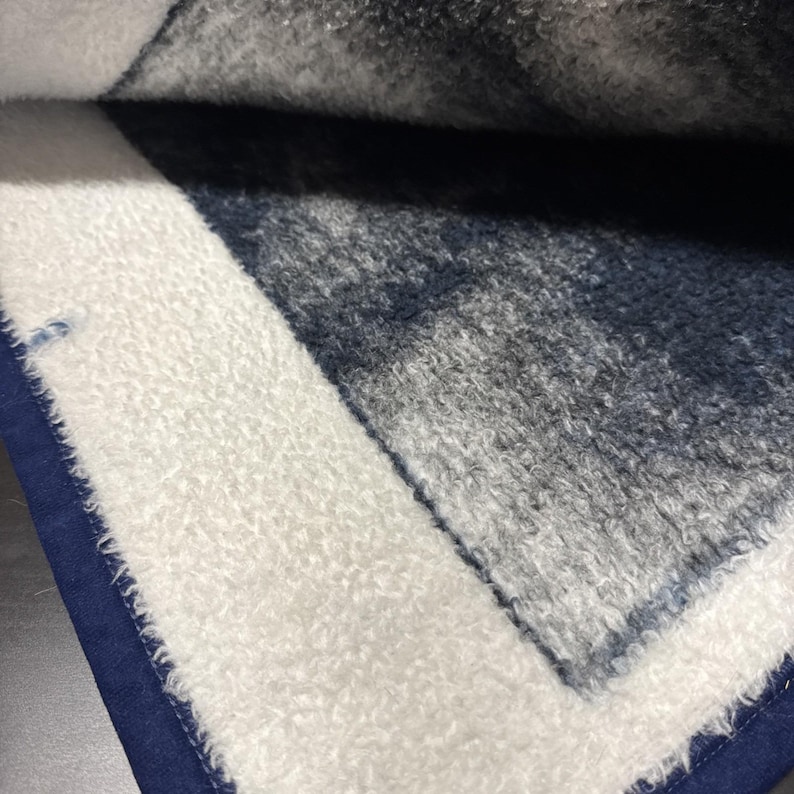 Puede incluir: Primer plano de una manta doblada con un borde blanco texturizado y un borde azul marino. El interior de la manta presenta una mezcla de tonos grises y azul marino, creando una apariencia suave y acogedora. La tela parece afelpada y c&oacute;moda.