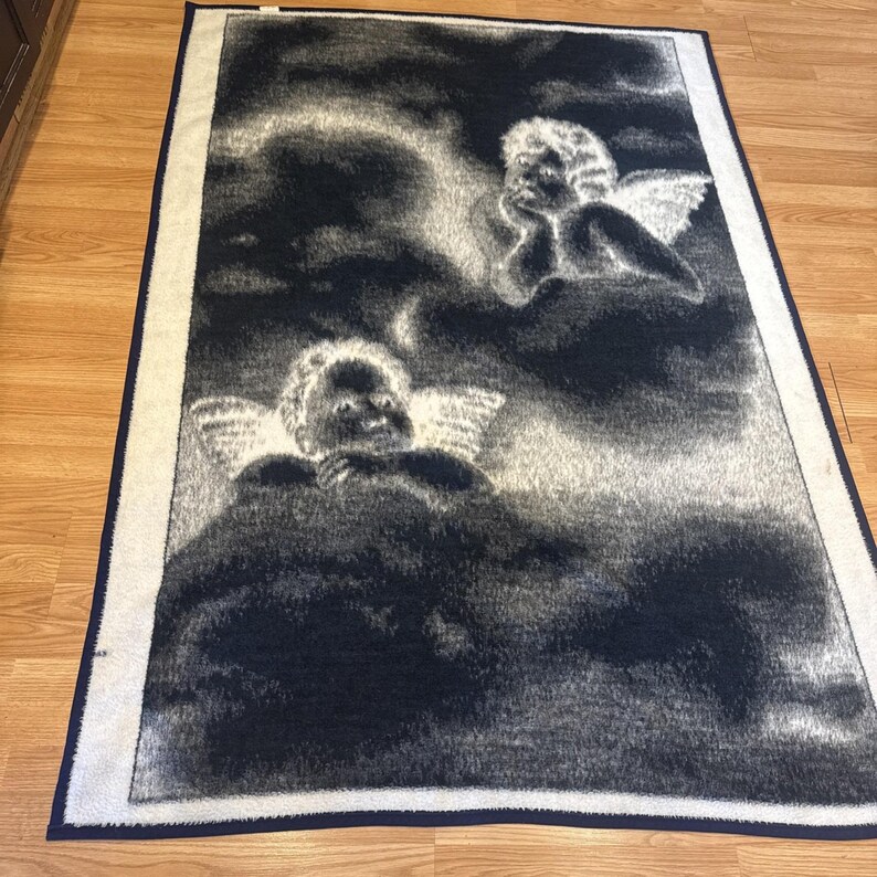 Puede incluir: Una manta rectangular en blanco y negro con un dise&ntilde;o celestial. La manta representa dos figuras de querubines con alas en medio de un fondo nublado. El borde es blanco con un ribete azul marino.