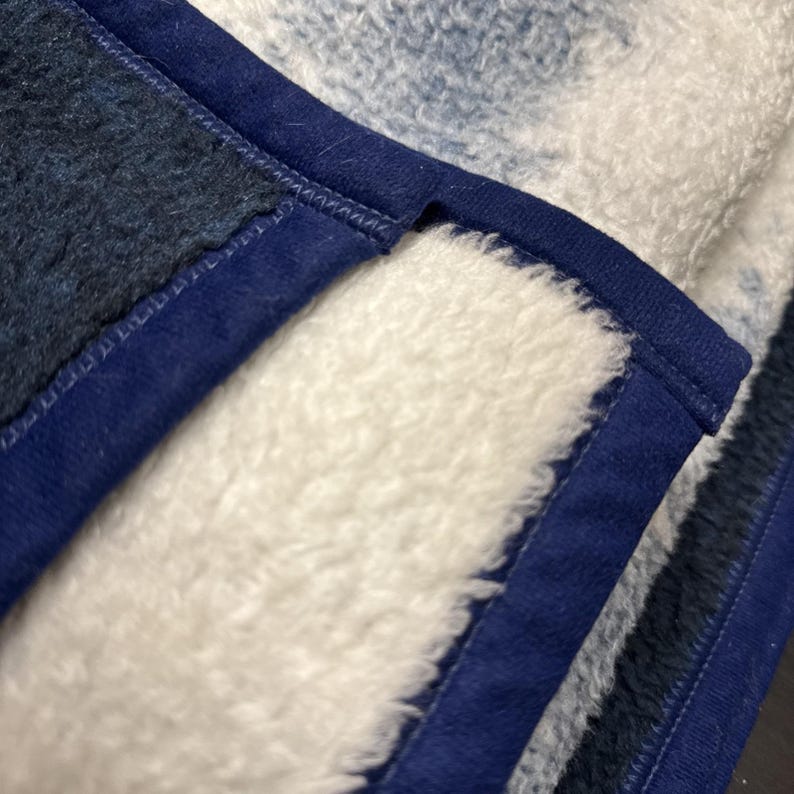 Puede incluir: Primer plano de una manta suave y afelpada con un estampado tie-dye blanco y azul. La manta tiene un borde azul oscuro y una superficie esponjosa y texturizada, perfecta para la comodidad.