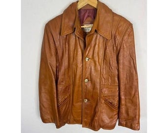 Peut inclure: Veste en cuir marron vintage avec un col et trois boutons dorés. La veste a deux poches avant et est suspendue à un cintre en bois. La veste est de couleur marron moyen.