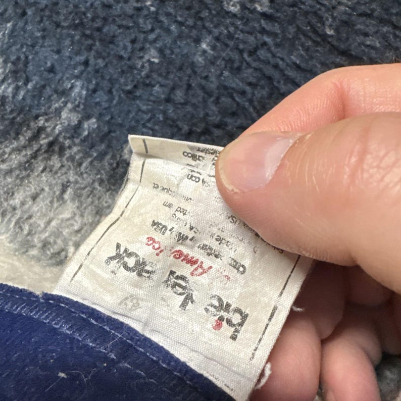 Puede incluir: Primer plano de una etiqueta de prenda. La etiqueta blanca tiene texto negro que dice "Big Joe Jack America". La etiqueta est&aacute; adherida a una tela azul. El fondo es una tela azul y gris texturizada.