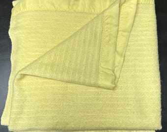 Peut inclure: Une couverture jaune pâle douce présentant un motif tricoté texturé sur toute sa surface. La couverture est finie avec une bordure en tissu satiné assorti de couleur jaune qui encadre les bords, offrant un aspect propre et uniforme.