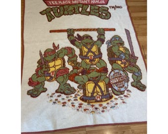 Peut inclure: Une couverture rectangulaire blanche ornée du logo Teenage Mutant Ninja Turtles en texte rouge et vert. Le motif représente quatre tortues vertes avec des plastrons jaunes, des masques rouges et des armes, incluant une plaque d'égout avec du texte.