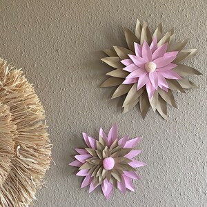 Peut inclure: Deux décorations murales en forme de fleurs en papier. Les fleurs ont un design en couches avec des pétales beiges et roses, et un petit centre en pompon rose. Les fleurs sont montées sur un mur texturé de couleur neutre.
