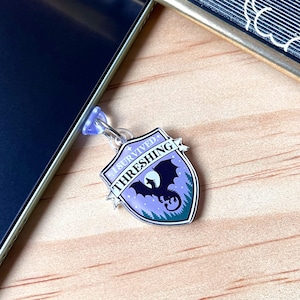 May include: A shield-shaped charm featuring a purple and teal gradient background with a black dragon silhouette and the text I SURVIVED THRESHING. The charm has a silver-toned metal border and is attached to a small, clear plastic suction cup.