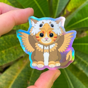 Könnte beinhalten: Ein holografischer Aufkleber mit einer Cartoon-Katze, die als Greif verkleidet ist. Die Katze hat orangefarbenes und weißes Fell und trägt ein braunes und beiges Kostüm mit Flügeln und einem vogelartigen Kopfschmuck. Der Aufkleber hat ein glänzendes, irisierendes Finish.