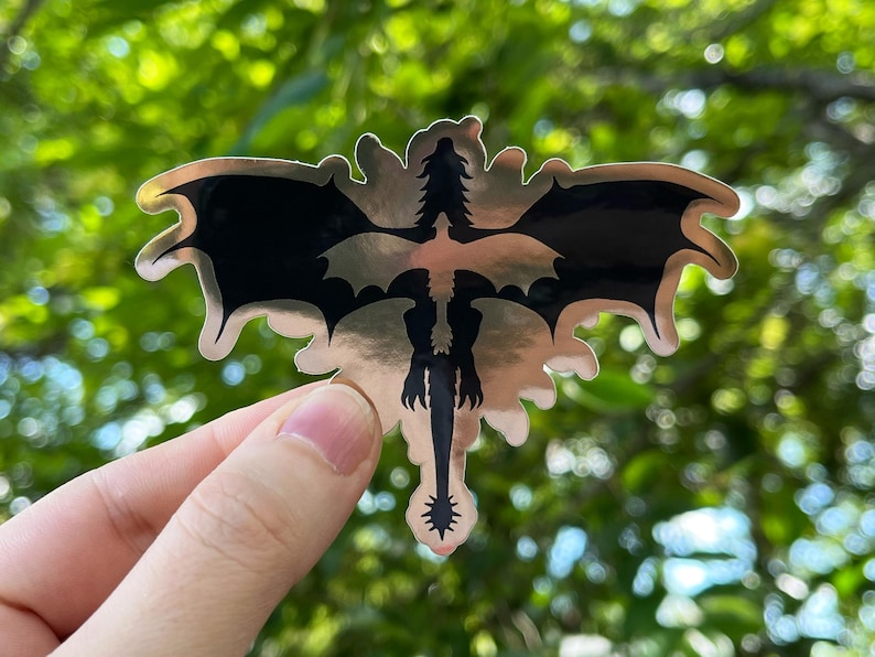 Puede incluir: Una pegatina met&aacute;lica con un dise&ntilde;o de drag&oacute;n estilizado. El drag&oacute;n es negro con alas extendidas y una larga cola, sobre un fondo plateado. La pegatina tiene un borde decorativo y ornamentado.