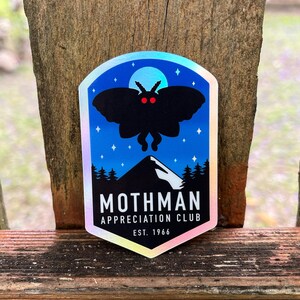 May include: A holographic sticker featuring a black silhouette of a moth with red eyes, set against a blue night sky with stars and a mountain range. The text reads "Mothman Appreciation Club, Est. 1966."