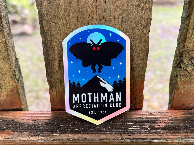 May include: A holographic sticker featuring a black silhouette of a moth with red eyes, set against a blue night sky with stars and a mountain range. The text reads "Mothman Appreciation Club, Est. 1966."