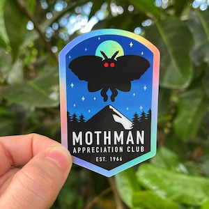 May include: A holographic sticker featuring a black silhouette of a moth with red eyes, above a mountain scene. The sticker reads "Mothman Appreciation Club" and "EST. 1966" against a blue night sky with stars and a moon.