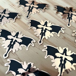 Puede incluir: Una colecci&oacute;n de pegatinas con forma de drag&oacute;n. Cada pegatina presenta una silueta de drag&oacute;n negro con alas extendidas y una larga cola, sobre un fondo de oro rosa met&aacute;lico. Las pegatinas est&aacute;n dispuestas sobre una superficie de madera clara.