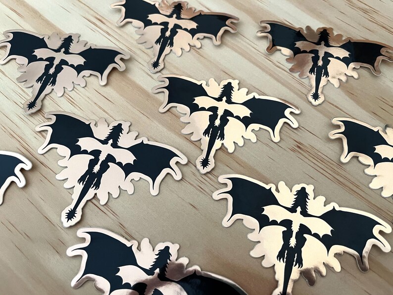 Puede incluir: Una colecci&oacute;n de pegatinas con forma de drag&oacute;n. Cada pegatina presenta una silueta de drag&oacute;n negro con alas extendidas y una larga cola, sobre un fondo de oro rosa met&aacute;lico. Las pegatinas est&aacute;n dispuestas sobre una superficie de madera clara.