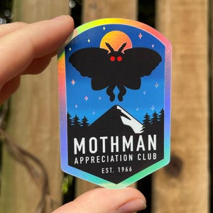 May include: A holographic sticker featuring a black silhouette of a moth with red eyes against a blue night sky with a mountain and trees. The text reads "Mothman Appreciation Club Est. 1966".